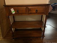 Front view showing the cabinet with two drawers and two shelves, wood stained medium brown with black ring pull handles. Visible wear and scratches.