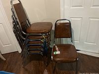 Photo showing 6 stackable cushioned metal chairs with chrome frames and brown vinyl cushioned seats and backs, stacked against a wall