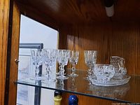 8 crystal wine glasses and crystal creamer and sugar bowl set on oval tray behind a glass cabinet door