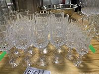 Photo showing a group of around 24 clear crystal glasses of different sizes arranged on a wooden table. The glasses have detailed cut patterns and long stems.