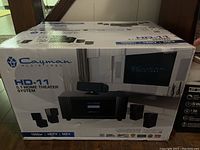 Photo of boxed Cayman Media Labs HD-11 5.1 home theater system showing front and side packaging with branding and product image.