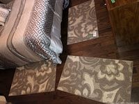 Three beige and white patterned floor mats arranged near a bed on a wooden floor, showing their size and wear