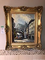 Full view of the oil painting in an ornate gold-colored frame depicting a street scene with buildings and people.