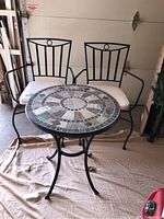 Full view of the outdoor metal table with mosaic stone top and two matching metal chairs with cream cushions, located in a garage.