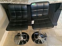 Pair of black vinyl adjustable stools with chrome bases placed side by side showing front views of seats and backs.