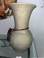 Side view of a large beige pottery vase with tassels on a glass shelf.