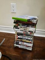 Stack of multiple Xbox, PlayStation 2, and 3 game cases and discs stacked on wooden floor showing titles such as NHL 07, NHL 2004, Sonic Heroes, WWE 13, and FIFA Soccer 08