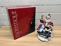 Santa Claus figure on sled next to original Clothtique Possible Dreams box with red background and tree graphic, photographed against a white brick wall.
