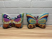 Two Coldwater Creek butterfly-shaped throw pillows side by side on a wooden surface against a white brick wall backdrop.