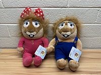 Front view of two stuffed gopher toys side by side, one in pink pajamas with polka dot bow, other in blue overalls, both with tags attached