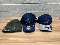 Three Dallas Cowboys hats: one knit beanie and two fitted caps, displayed on a wooden table against a cinderblock wall.