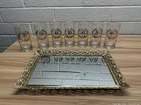 Eight 50th anniversary decorated glasses arranged in front of an ornate gold framed mirror serving tray on a wooden surface against a brick wall background.