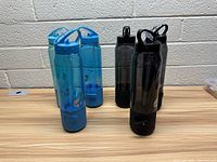 Blue and black Bluetooth speaker water bottles arranged in two rows on a table in front of a brick wall.
