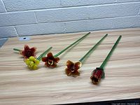 Four glass flowers on a wooden surface, green stems, colorful blooms in shades of red, orange, and yellow. One bloom detached.