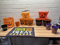 Photo of all Halloween buckets arranged on table showing orange and black buckets with various jack-o'-lantern and cat faces plus purple small buckets and one fabric bucket with googly eyes.