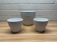Three ceramic popcorn bowls, white with embossed 'POPCORN' text, arranged in a row on a wooden surface against a brick wall.