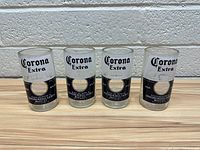Front view showing four clear shot glasses with Corona Extra branding spaced evenly in a row on a wooden surface against a white textured wall.