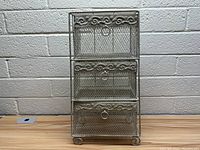 Front view of the small metal basket organizer showing three mesh baskets with decorative scroll designs and ring pull handles, set against a gray brick wall.