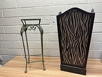 Side view of small green metal plant stand next to black decorative trash can with zebra fabric sides on wooden surface with white brick wall background.