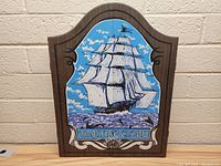 Front of closed wooden wall mount dart board cabinet showing a decorative painting of a tall sailing ship on the doors titled 'The Flying Cloud'