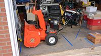 Full side view of orange housing, auger, wheels, handlebar and engine
