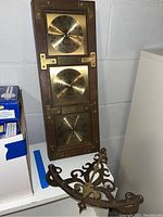 Full view of the wall-mounted weather station with three brass dials mounted on wood, alongside the separate brass sundial on a white shelf.