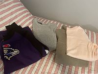 Photo of folded mens shirts and sweaters on bed showing a purple Baltimore Ravens shirt, a black shirt, a gray sweater, a beige sweater, and a light pink womens long sleeve t-shirt.