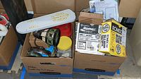Two boxes filled with electrical and houseware supplies including Romex wire in labeled boxes, a ceiling fan blade with white and gold detailing, and a wall mount lamp with stained glass shade.