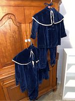 Two navy blue velvet children's outfits hanging on an armoire. One outfit has a cape with white trim.