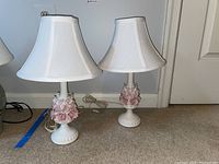 Two matching white ceramic lamps with pink flower decorations at the base, placed side by side on the floor with cords attached.