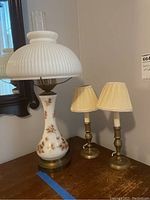 Porcelain lamp with white ribbed glass shade and two smaller brass candle lamps with cream pleated shades on wooden table.