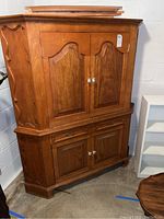 Full front view of the two-piece corner media cabinet showing the removable top section and bottom cabinet with doors and drawer. Warm medium brown wood finish with white round knobs.
