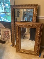 Two gold framed mirrors leaning against a wall, showing ornate carved gold frames with floral and scroll patterns. The mirrors reflect a room interior.