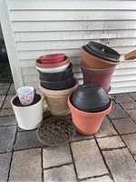 Photo showing a collection of stacked plastic and terracotta gardening pots, a small white ceramic planter, and a round decorative metal grate on a stone tile floor.