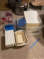 Photo showing a variety of plastic storage bins with lids (white, blue, and red) and nested storage baskets piled on carpet.
