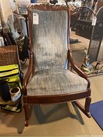 Front view of the wood framed rocking chair showing upholstered blue-gray fabric seat and back, detailed carved wood top, and rocking base.