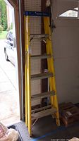 Full view of yellow Werner fiberglass ladder, six feet tall, standing in garage near garage door. Shows some dirt and stains from use.