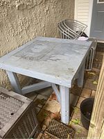 Custom built gray wooden square table with weathered surface showing peeling paint and staining, stable legs with cross braces, pictured outdoors on brick patio alongside chairs and other items.