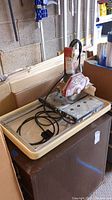 MK tile cutter on metal base with beige plastic collection tub underneath, showing the entire setup from a distance.