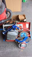 Top view of a toolbox containing various hand tools including knee pads, red caulk gun, metal saw frame, and circular cutting discs taped together.