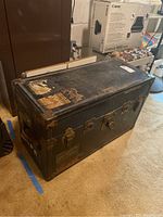 Closed view of vintage black travel trunk with metal latches and corner protectors, showing aged exterior and peeling labels.