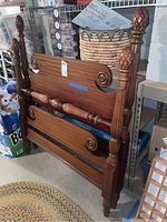 Wooden twin bed frame pieces showing decorative rounded posts with pineapple finials and scrollwork on the boards. Located in basement with shelving and other household items visible around.