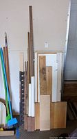 Photo showing multiple spare wood pieces leaning against wall, including shelving boards and thin pieces of wood, two white PVC pipes and a plastic paver edging.