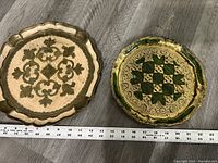 Top-down view of two round vintage decorative trays with gold, brown, and green patterns and scalloped edges, placed on wood surface with measuring tape showing about 12 inches diameter.