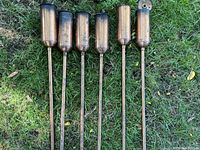 Six solid copper outdoor tiki torches laid on grass, showing their cylindrical fuel reservoirs and long poles.