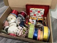 Wide view of box filled with various ribbons, pom pom makers, and craft supplies showing color variety and quantity.