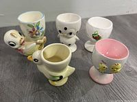 Five vintage ceramic egg cups with duck and chick designs shown from angled side view; includes detailed view of designs and shapes.