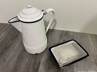 Photo showing the white enamel coffee pot with black trim and matching rectangular enamel tray on a floor surface.