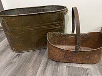 Side view of large antique copper boiler and oval copper basket with handle, both showing aged patina.