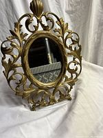 Front angle of vintage oval vanity mirror with gold gilt metal ornate frame showing leaf and scroll design.